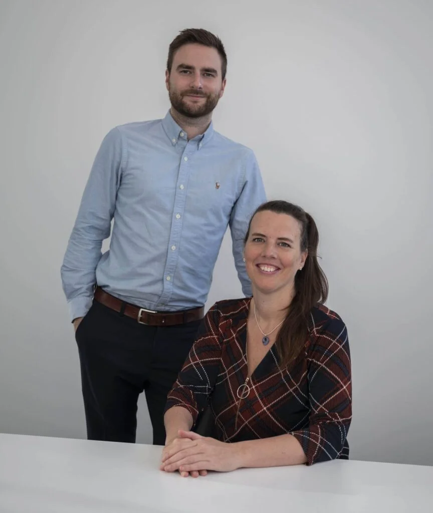 Een man in een blauw shirt staat met zijn handen in de zakken achter een zittende vrouw in een jurk met patroon aan een witte tafel. Beiden lachen tegen een effen grijze achtergrond.