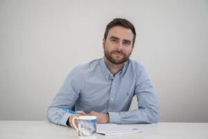 Een man met een baard en een blauw shirt zit achter een bureau, met een pen in zijn hand. Voor hem ligt een notitieboekje en er staat een mok in de buurt.