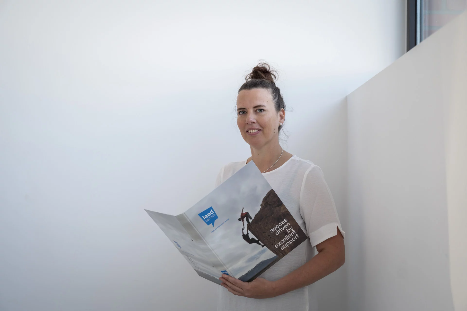 Een persoon in een wit overhemd staat bij een muur en houdt een opengeslagen brochure vast met een afbeelding van een bergbeklimmer op de omslag.