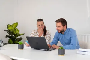 Twee mensen zitten aan een wit bureau, werken samen aan een laptop, met potplanten en notitieboekjes op tafel in een lichte kantooromgeving.