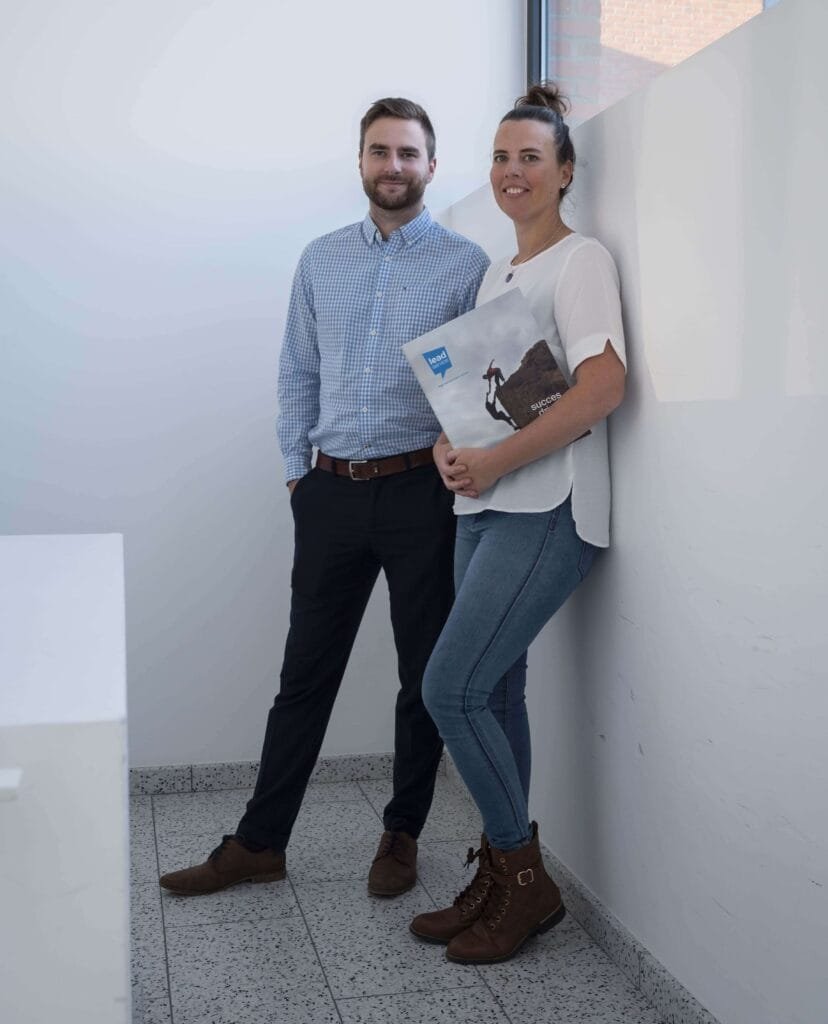 Een man in een geruit hemd en donkere broek staat naast een vrouw in een witte blouse en spijkerbroek met een map in de hand. Beiden poseren in een fel verlichte gang, mogelijk discussiërend over hoe te vzw oprichten.