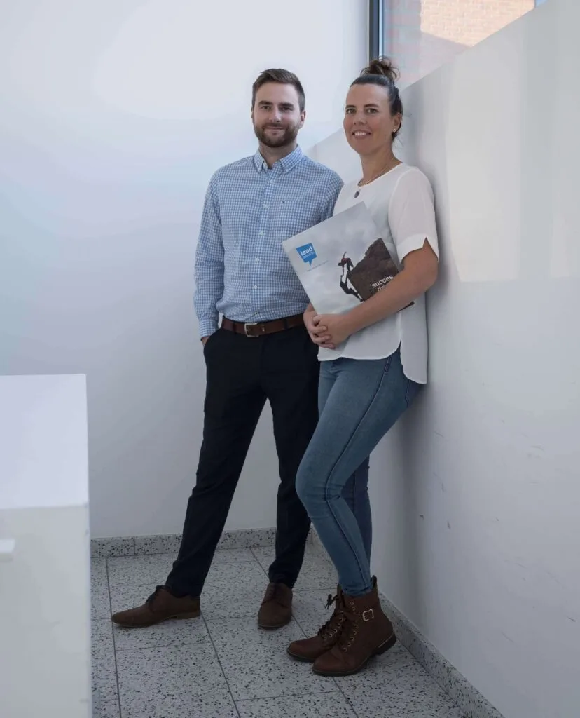 Een man in een geruit hemd en donkere broek staat naast een vrouw in een witte blouse en spijkerbroek met een map in de hand. Beiden poseren in een fel verlichte gang, mogelijk discussiërend over hoe te vzw oprichten.